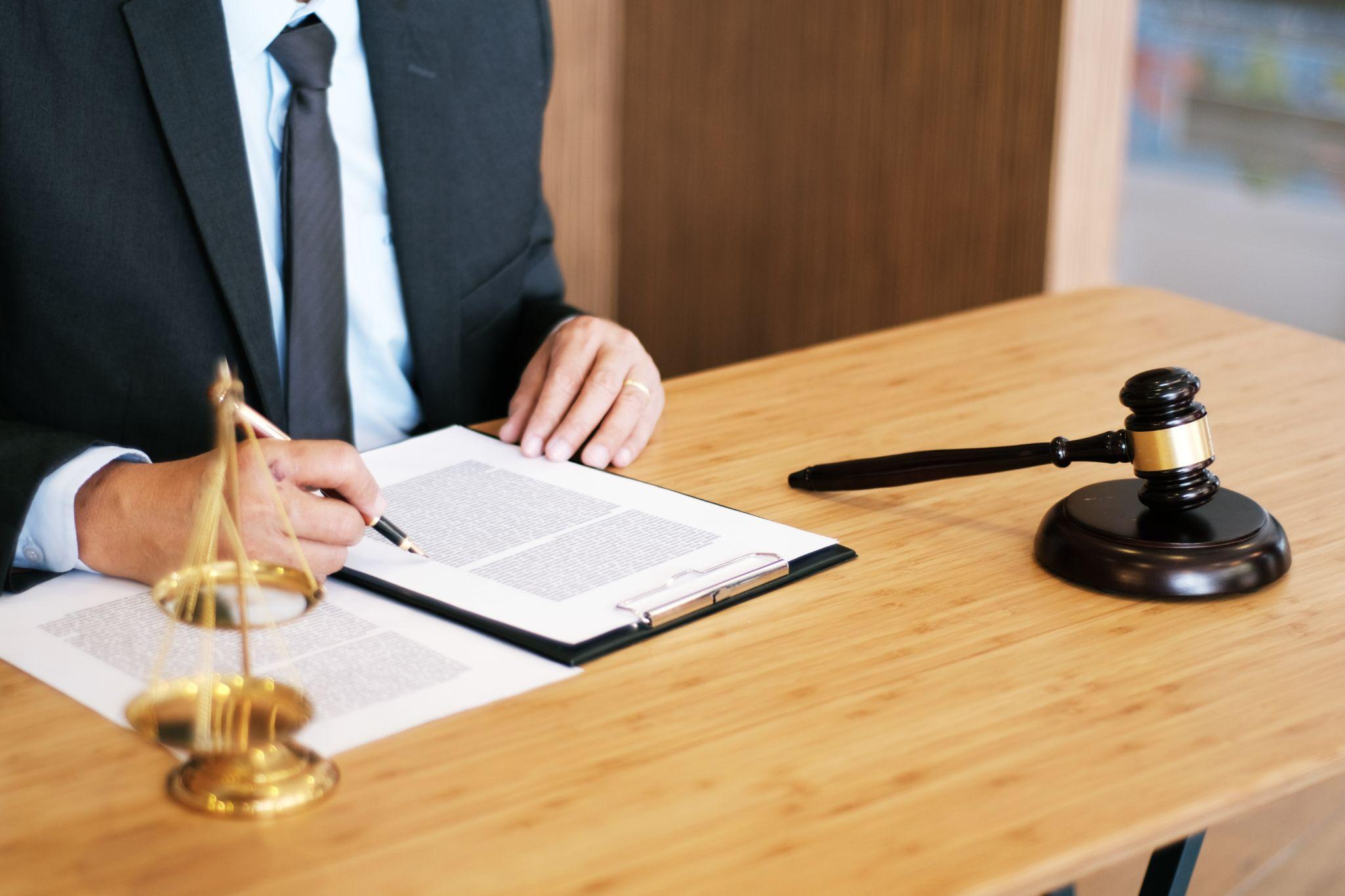 Lawyer in suit reviewing documents at a wooden desk with a gavel and scales of justice nearby.