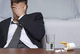 A man in a suit sitting on a bed next to a glass of pills, indicating possible addiction issues.