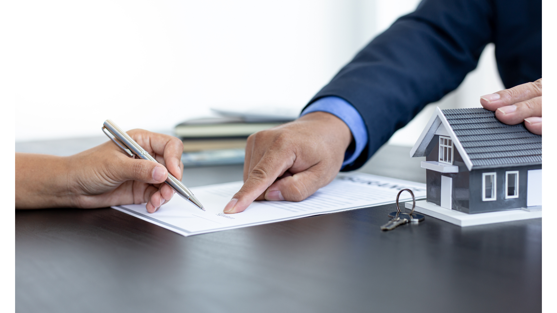 Two individuals at a table during a real estate transaction rwith one person pointing to a document, likely a contract, for the other to sign, with a small model