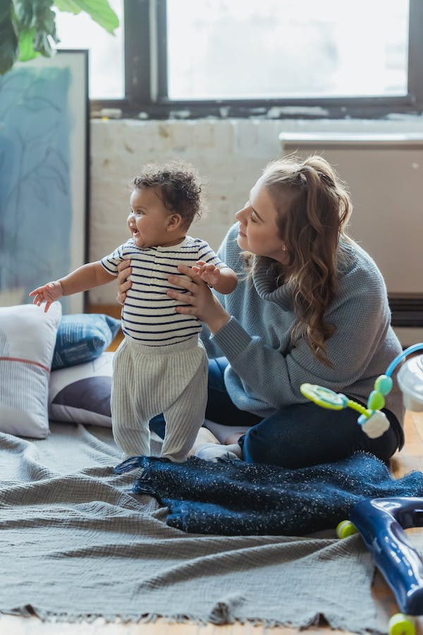 A woman helps a baby stand up on a play mat indoors.