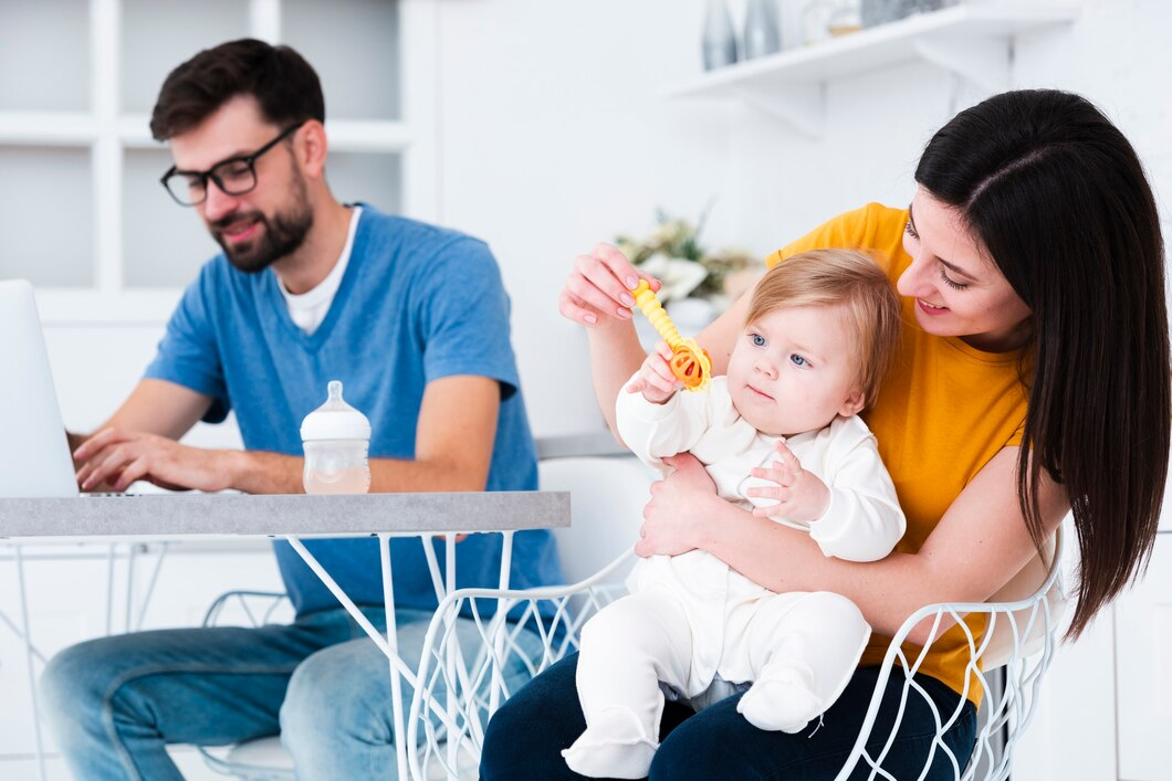 A mother holding a toddler plays with a toy while a father works on a laptop in a bright kitchen setting.