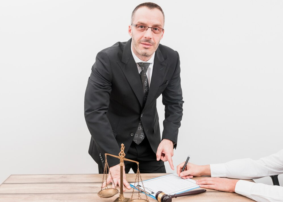 Man in a suit with glasses sitting at a table, about to sign a document with a small scale of justice in the foreground.