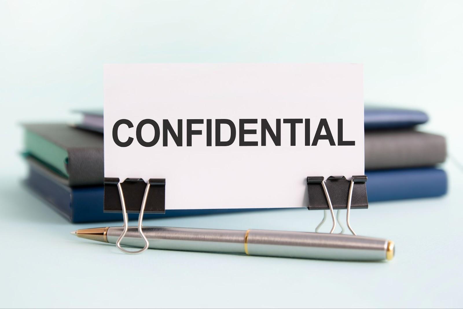 A card labeled "confidential" clipped to a pen in front of stacked notebooks