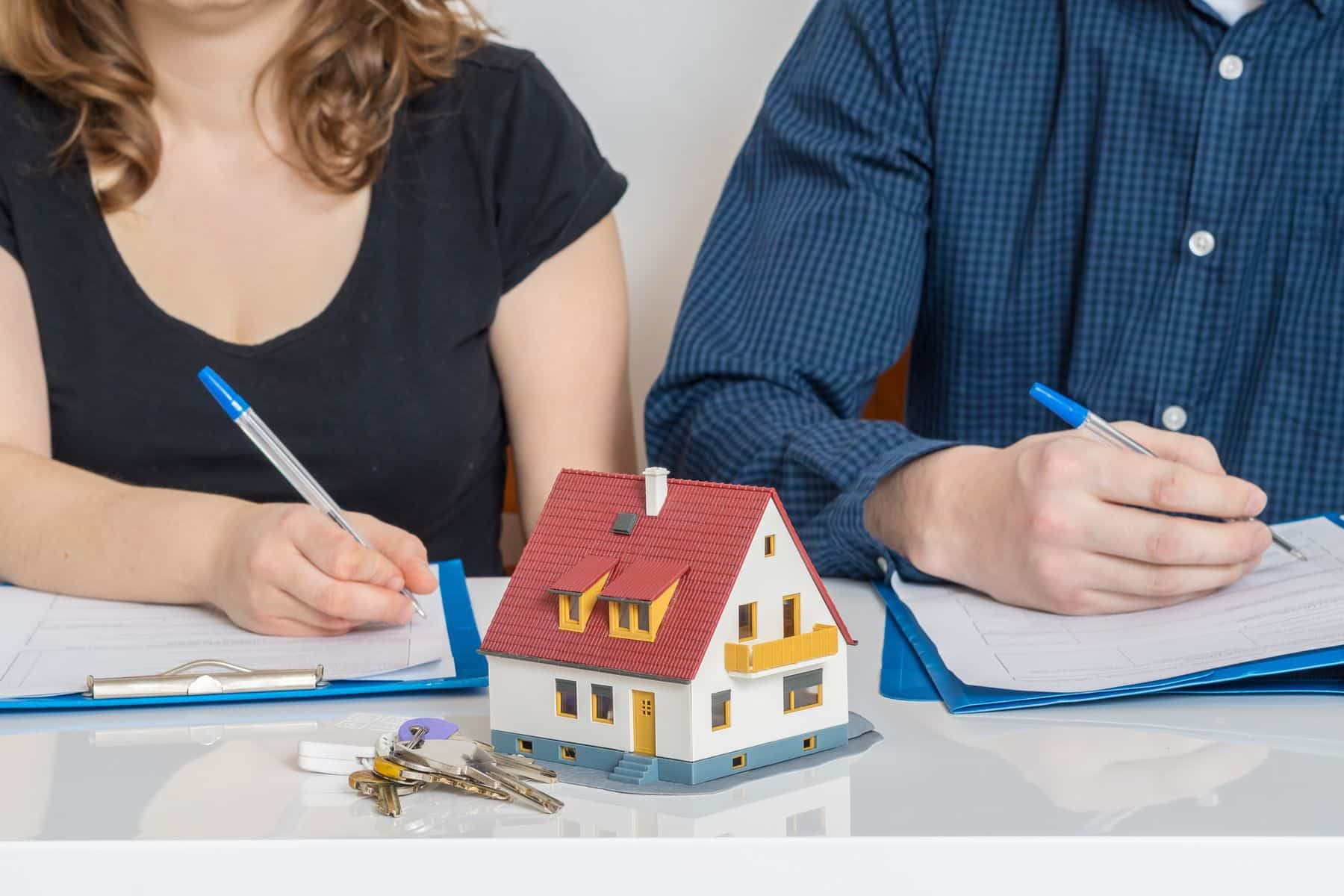 Two individuals with paperwork at a table featuring a model house and keys