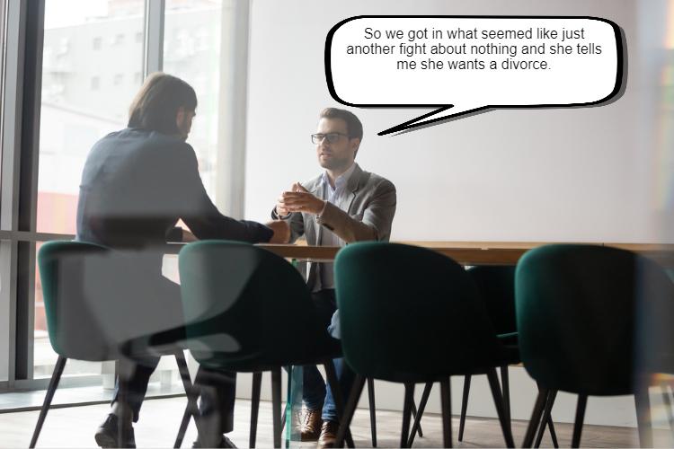 Two people having a serious conversation at a table, one gesturing while talking, in a brightly lit office setting.