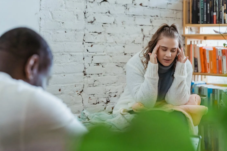 A woman in a white cardigan sitting on a green couch, smiling and talking to a man, who is seen blurred in the foreground, in a cozy room with a white brick wall and booksh