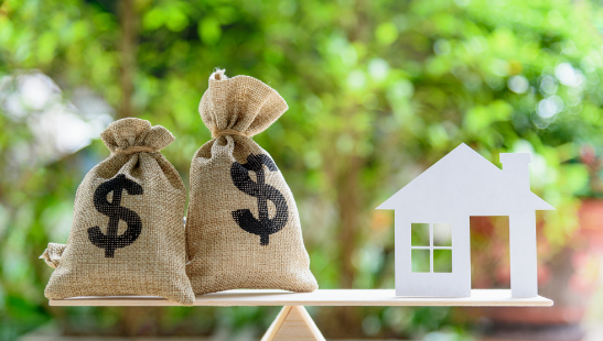 Two burlap bags with dollar signs and a white house cutout on a balanced beam