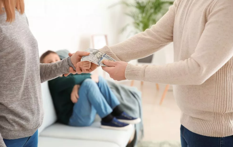 Two people exchanging money in a room while another person watches from a couch in the background.