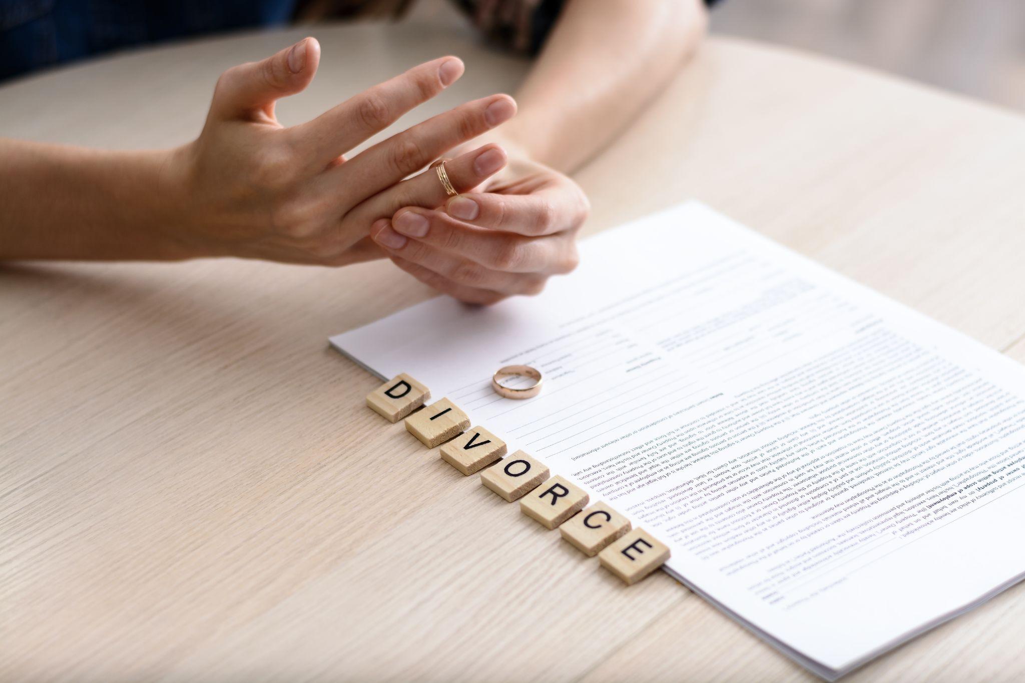 A person removing a wedding ring with the word "divorce" spelled out in tiles on a table next to a document.
