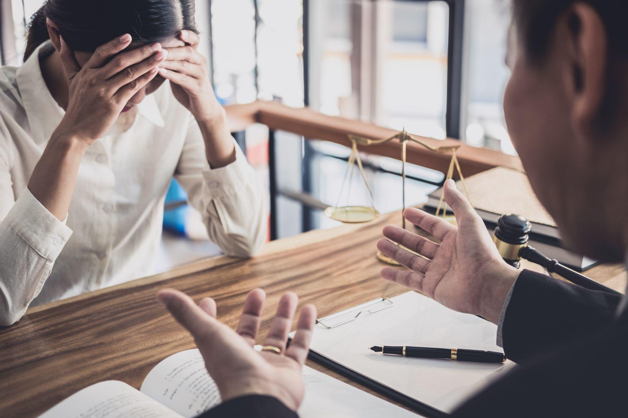 A woman appears distressed covering her face with her hands in a legal office, while a male lawyer gestures while speaking. documents and a gavel are visible on the table.