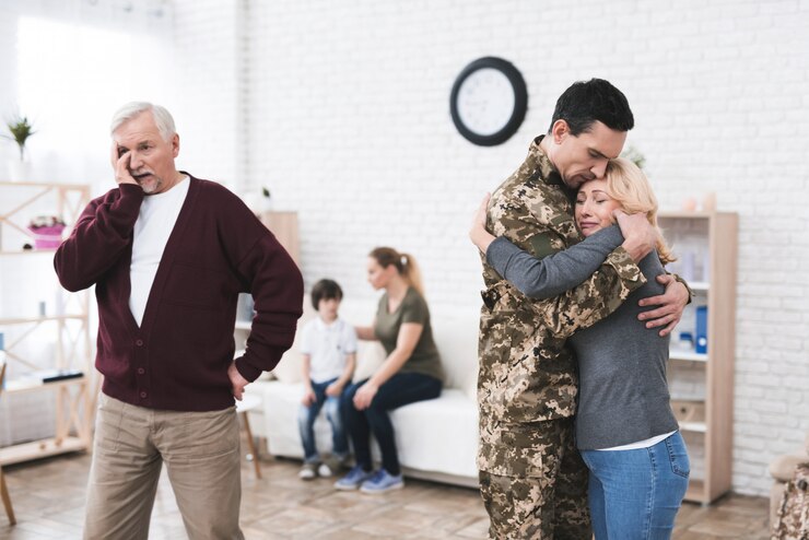 A soldier embracing a tearful woman, as an older man watches on a phone, with a family sitting in the background in a home setting.