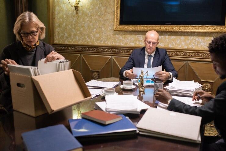Three business professionals review documents at a table filled with files and teacups in an ornate room.