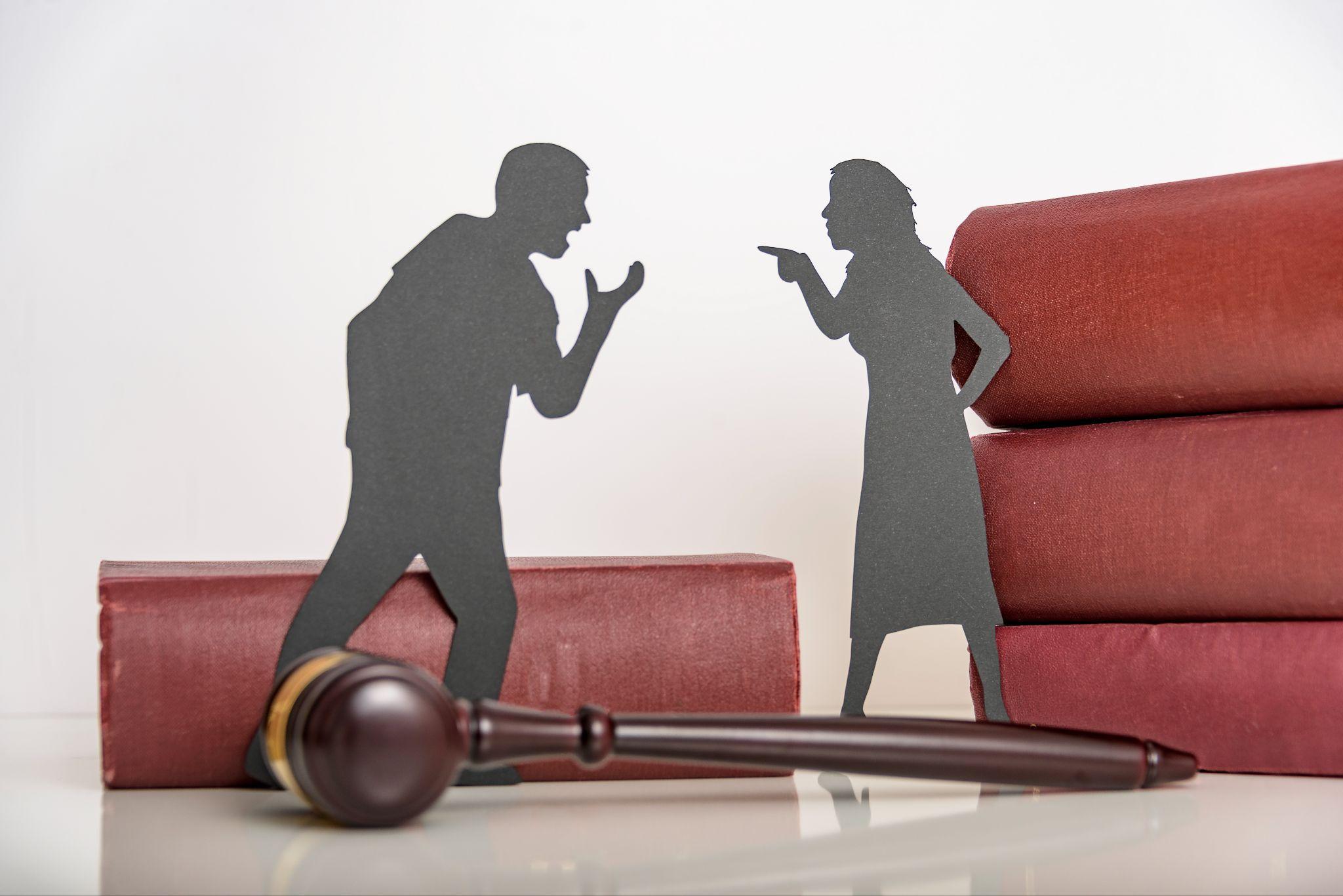 Paper cutouts of a man and a woman arguing next to a stack of red books and a wooden gavel on a white surface.