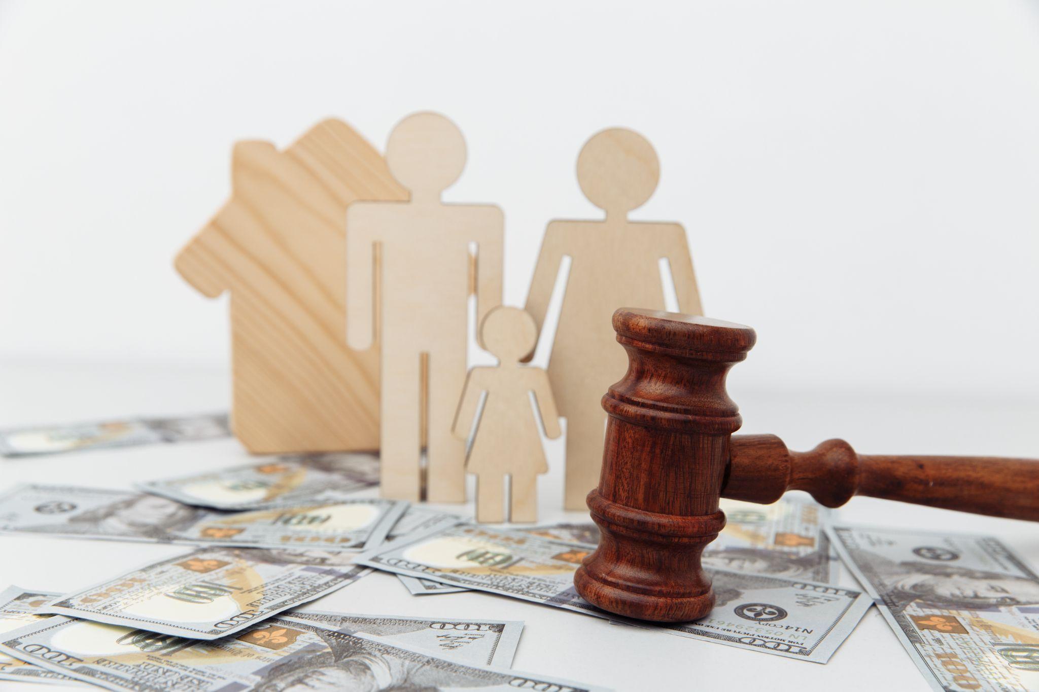 Wooden figures of a family and a house, a gavel, and scattered dollar bills on a table, symbolizing legal and financial aspects of family and property.