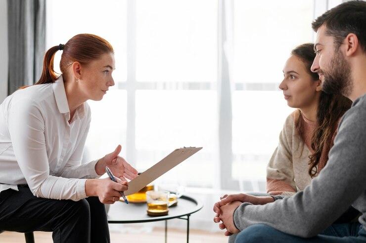 A woman holding a clipboard appears to be counseling a couple sitting opposite her on a couch. They are engaged in conversation in a well-lit room.