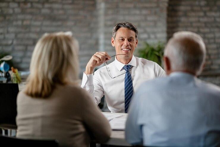 A man in a white shirt and blue tie is smiling while holding his glasses. He sits across from two people with their backs to the camera in an office setting,