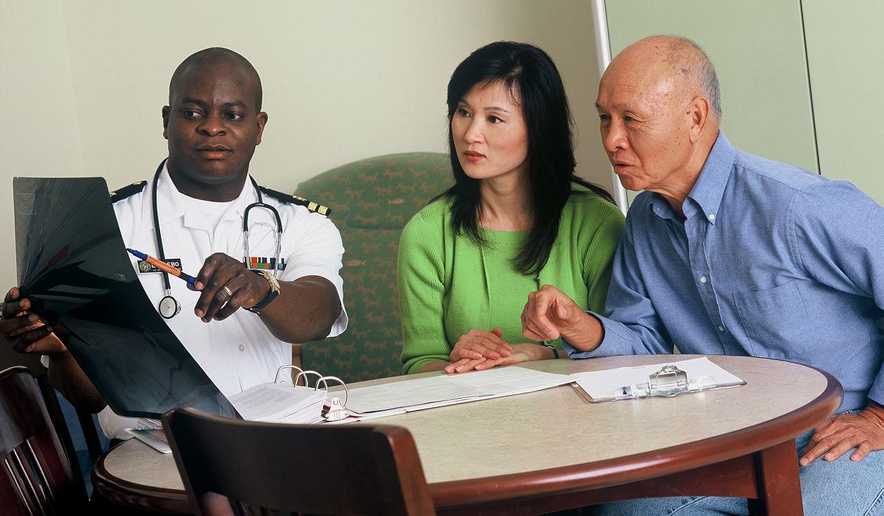 A medical professional shows an X-ray to two people sitting at a table during a consultation. One person points to the X-ray while the others listen attentively.
