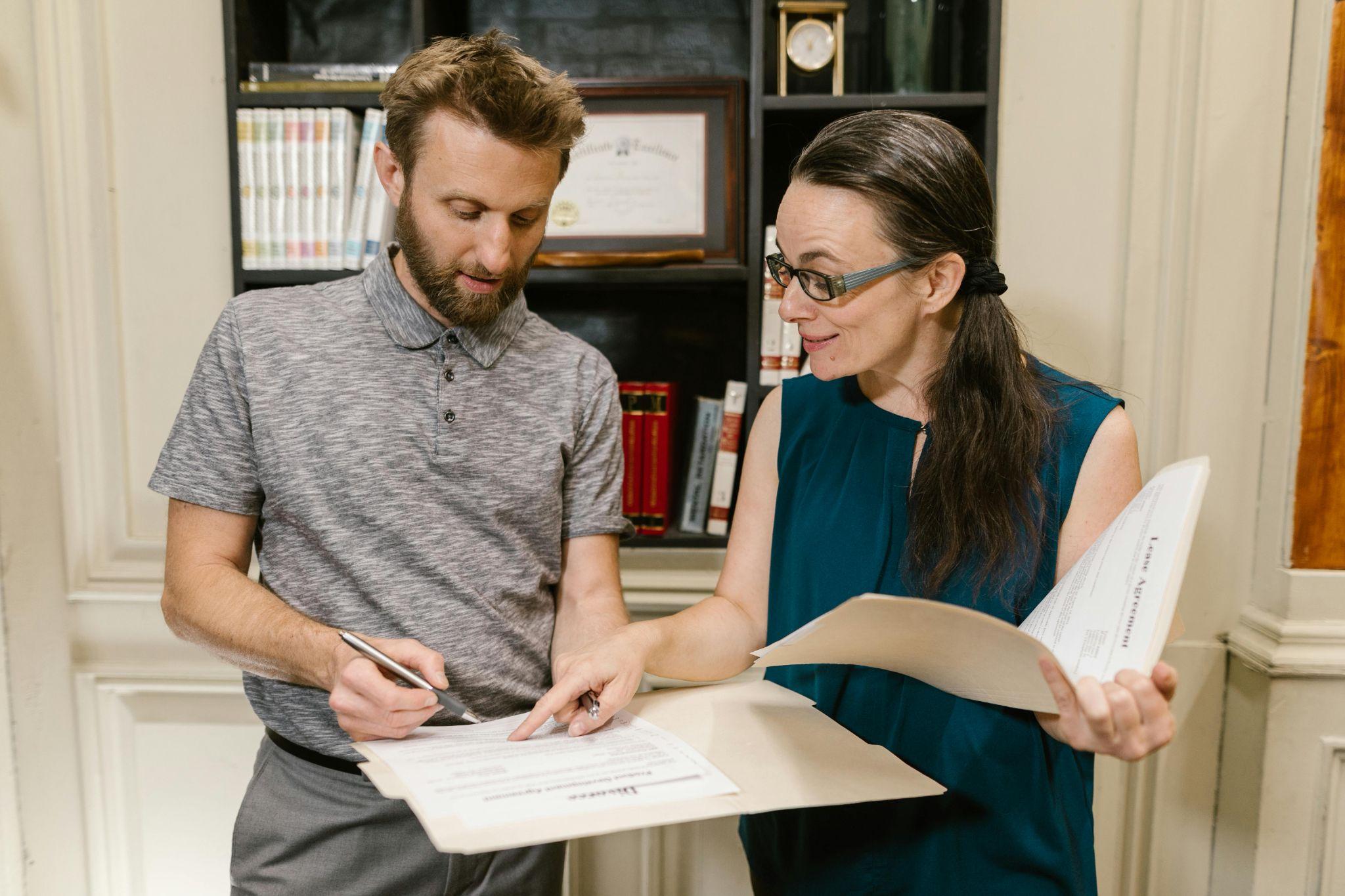 Two people are discussing paperwork in an office setting. One is holding a folder and pointing at a document while the other is writing on the paper.