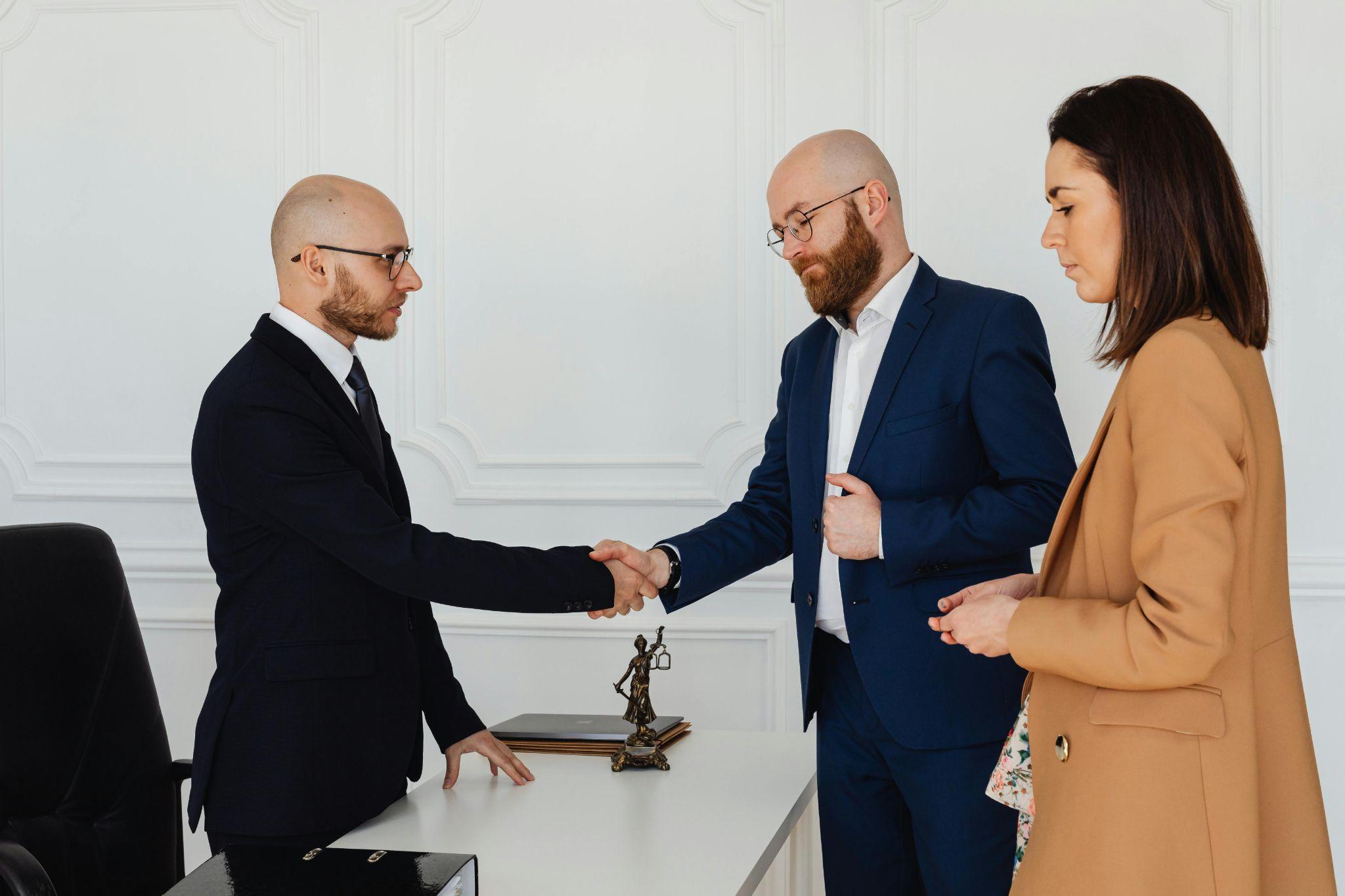 Two men in suits are shaking hands across a desk, while a woman in a tan coat stands beside them holding a pen. A statue of Lady Justice is on the desk.