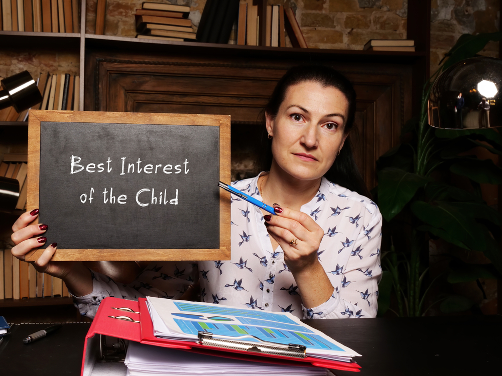 A woman holding a chalkboard sign that reads "Best Interest of the Child" while sitting at a desk with documents and a pen.