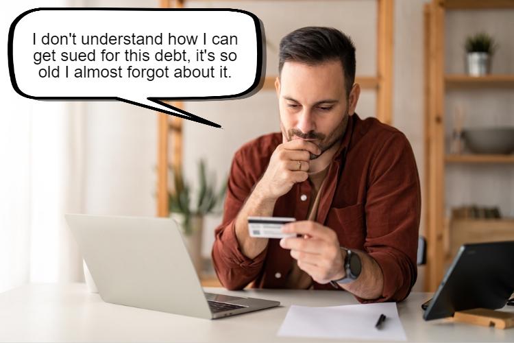 A man sits at a desk looking at a credit card with a puzzled expression. A speech bubble shows him saying, "I don’t understand how I can get sued for this debt, it's so old I almost forgot about it. When does the statute of limitations start for debt?