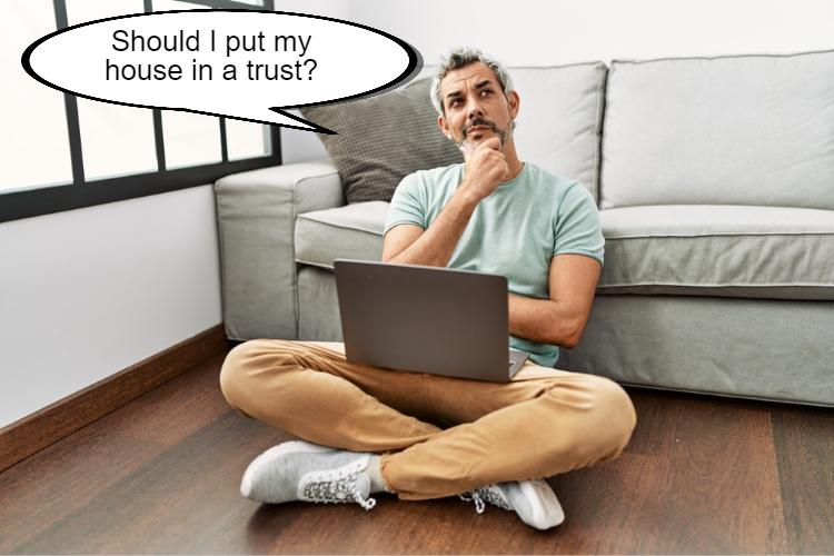 A man sitting on the floor with a laptop on his lap, looking thoughtful, with a speech bubble saying, "Should I put my house in a Texas Trust?