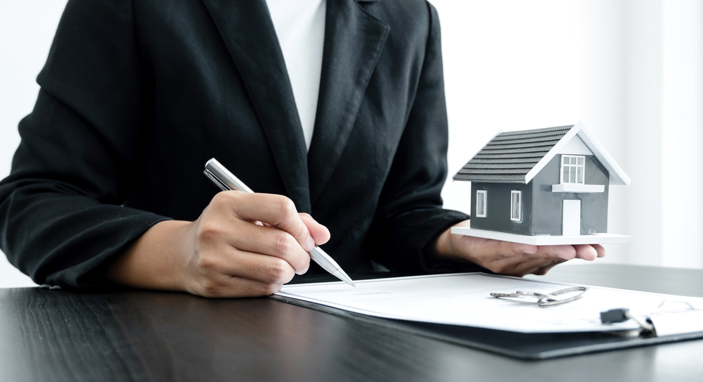 A person in a suit holding a small house model in one hand and signing a document with the other, while a set of keys rests on the table.
