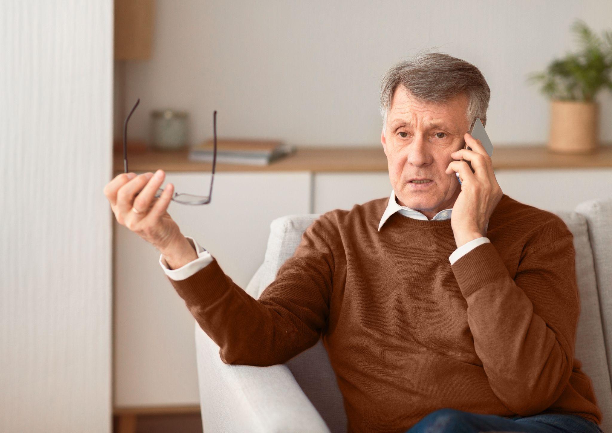 An older man sits on the couch, eyeglasses in one hand and smartphone in the other, discussing how a will safeguards his Texas estate and pondering over the implications of a no contest clause. His expression is one of deep concern.