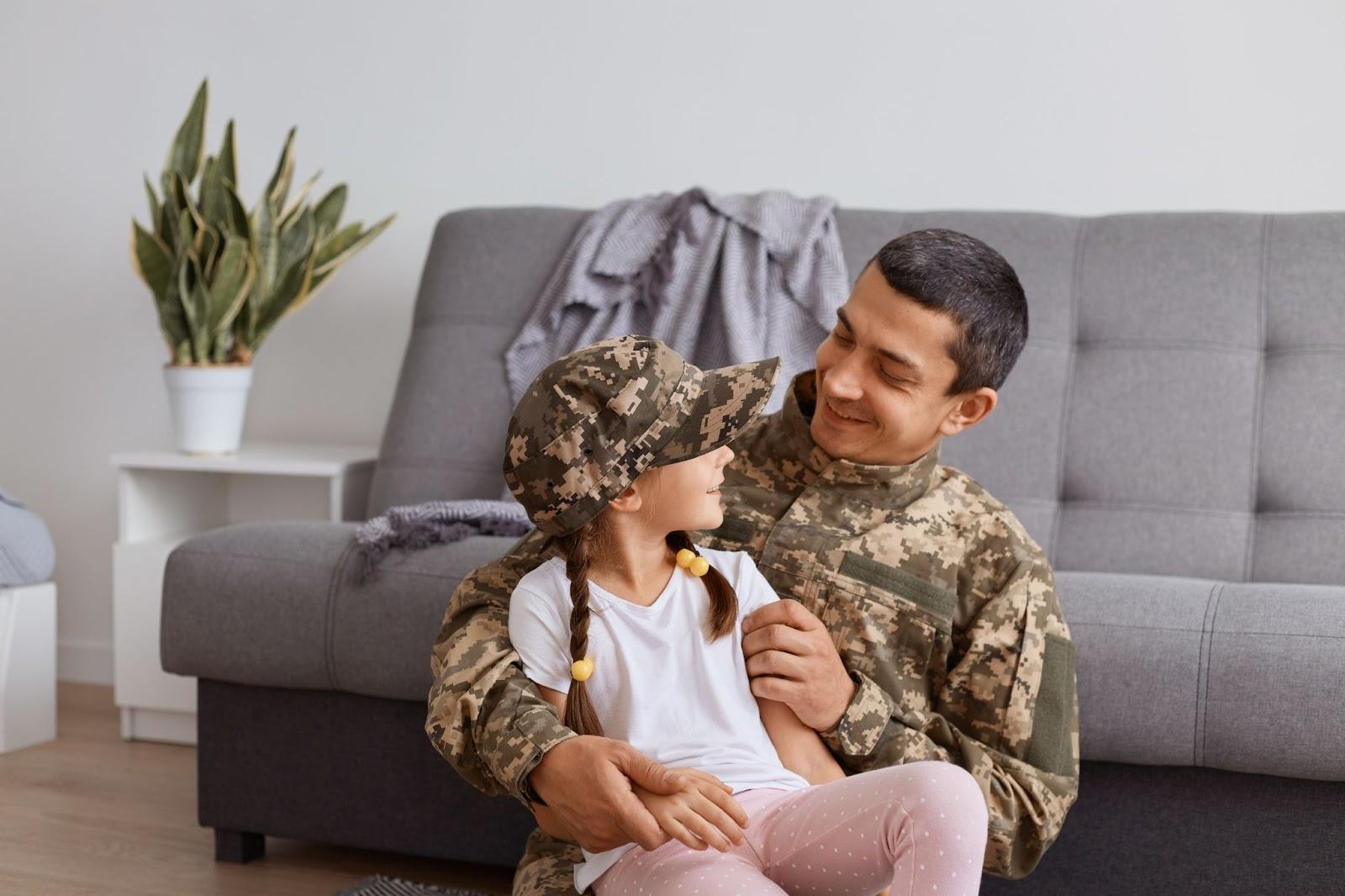 A soldier in a camouflage uniform smiles at a young girl wearing a matching cap, sitting on his lap in a cozy living room, reflecting on the challenges ahead with his military divorce lawyer.