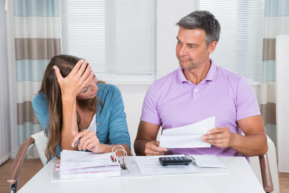 Man and women sitting next to each other at a table with a binder filled with papers open, the man is holding a paper and the women has her hand on her head 